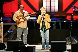 A blonde woman wearing a tan jacket and blue pants, singing into a microphone on a stage.  A man playing a guitar is visible in the background.