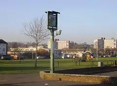 A photograph of an urban park with houses and flats in the distance.  The park large, and mostly grass.  A football goal is in the centre of the park, and a pub sign is in the foreground.