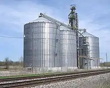 Three tall cylindrical metallic structures, reflecting sunlight, with slightly conical tops connected by tubing, rise behind railroad tracks in the foreground.