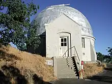 Stairs leading up to a small white building with a domed silver roof next to a tree.