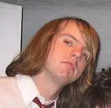 A man with long blonde hair in a white collared shirt faces the camera, head tilted to the right side of the frame and slightly upturned.