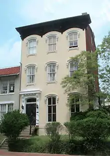 A yellow three-story townhouse with steps in front and a small lawn with shrubs and trees.