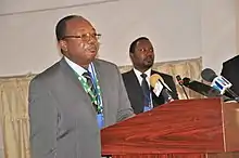 Man with glasses, standing at a podium with microphones