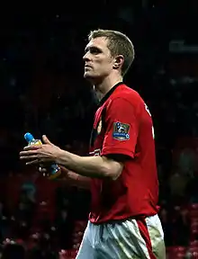 A white man with light hair standing with his hands on his hips. He is wearing a red football shirt.