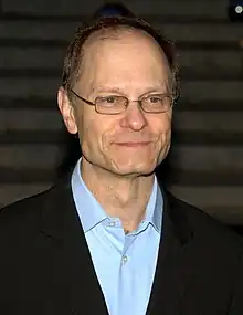 A man in his early fifties looking to the left of the camera. He is wearing thin rectangular glasses and a black blazer with a light-colored shirt.