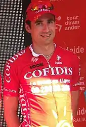 A man of about thirty wearing a mostly red cycling jersey with white trim standing with his hands behind his back.