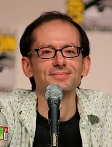 A closeup of a man in front of a microphone. He has a receding hairline and wears dark-framed glasses.