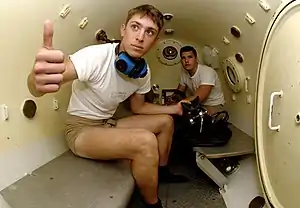 Photograph of the cramped interior of a cylinder containing two benches and two diver trainees