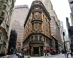 The facade of 56 Beaver Street as seen from the corner of Beaver and South William Streets. The facade is made of light orange and dark brown brick. There is a sharp curve in the center of the facade, between two streets on either side. A dark brown skyscraper is in the background.