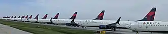 Many in line airplanes with the Delta Air Lines logo on the tail, parked on pavement behind a fence.