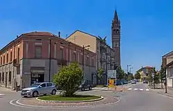 A small silver hatchback going around a roundabout in the foreground with a two-story tile-roofed building behind it. On the left side of a street leading into the roundabout from the background is brick campanile rising against a clear blue sky. There are signs in Italian and a crosswalk just behind the roundabout.