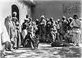 Woman in 19th century British dress stood amongst people in Afghanistan dress. She is positioned beside a doorway, and is examining the eye of a child.