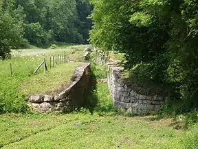 A view up three derelict canal locks. The canal is dry and the channel overgrown with grass and weeds. Deciduous woodland is in the distance