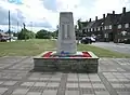 Gable end terraced housing with steep, tall hipped roofs by the war memorial with tessellating paving, square green and trees in the background.