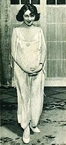 A young smiling white woman with bobbed dark hair, wearing a light-colored long gown