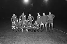 Black and white photo of a football team in kit, with a black sky in the background. Five players are crouched down, and six are standing.