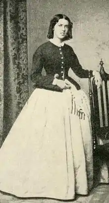 A young white woman, standing, wearing a fitted long-sleeved bodice and a long voluminous skirt.