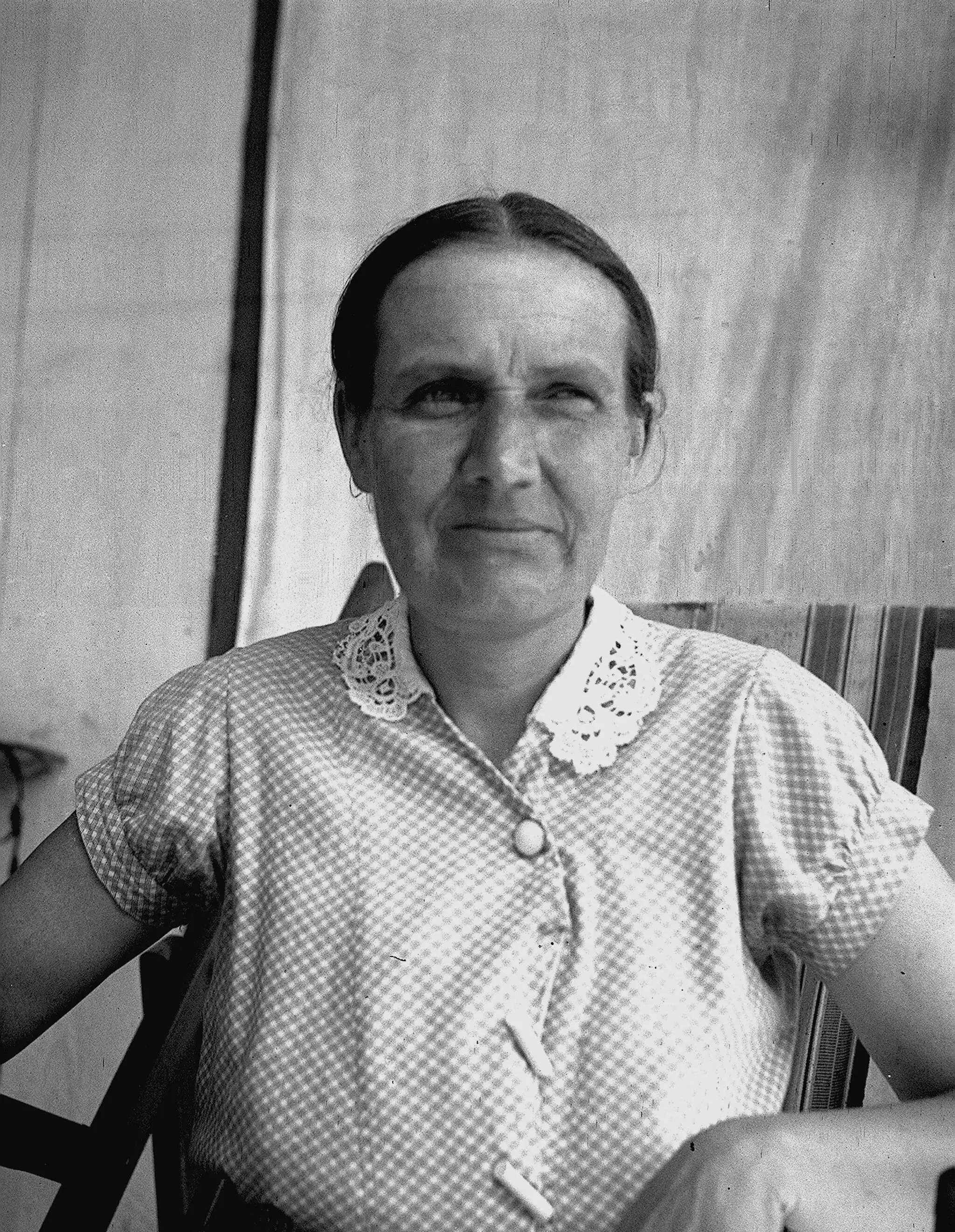 A severe, unsmiling 48-year-old woman in gingham short-sleeved blouse, with lace at the neck and two toggle buttons on the front.