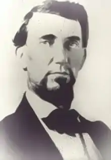 Photo shows a black-haired bearded man in a black civilian suit.