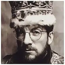 A close-up headshot of a man wearing glasses and a giant bejeweled crown on his head.