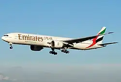 A mostly white Boeing 777, with some red, green and black markings, of Emirates, in flight, facing left.