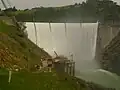 Englebright Dam during spring floods.  Water is spilling over the dam's crest.  Narrows II Powerhouse is visible at the base of the dam.