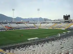 A semi-panoramic view of the stadium. The seats are white and black, and shows the club badge in the seats. The roof of the stand is supported by a cantilever structure, in a "buried" construction.