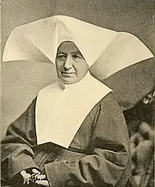 An older white woman in the habit of a religious sister, with a wide white wimple. She is holding rosary beads.