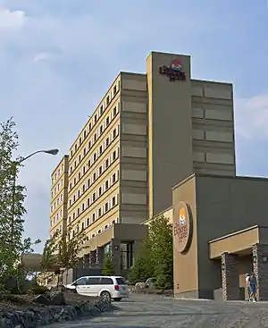 A beige and tan concrete high-rise seen from the base of an upwardly sloping gravel driveway curving around a planted area. On the building, and the side of a pavilion projecting to front right, are circular decorative signs identifying it as the Explorer Hotel.