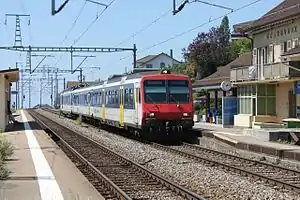 White and blue train with red front on a double-tracked railway line surrounded by platforms