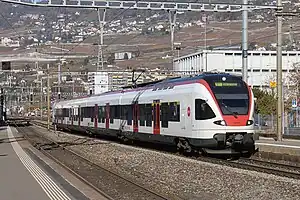 White train on tracks next to platform with a hillside rising up behind