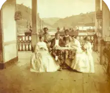 a man and three women seated around a table with two servants standing in the background