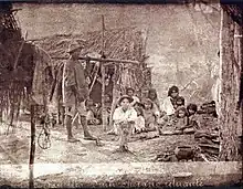 An old photograph depicting a group of poorly clad people in fronto of a small hut constructed of small tree branches