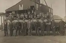 In a black and white photograph, group of 26 young to middle-aged men wearing suits and flat caps stand in two rows. Behind them is a large, industrial shed, and in the distance the roof of a house can be seen.