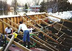 Several men setting up a wooden dike supported by a wooden frame against a river. A sandbag dike can be seen at the right of the image against the river. Several trees and homes can be seen in the background.