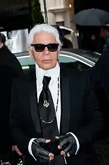 Karl Lagerfeld, who is an older man, wearing a black formal jacket, black gloves, a black tie, and black sunglasses, standing near a Fendi store.