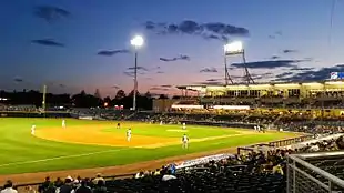 A view of the green baseball field from the third base side seats showing men in white baseball uniforms playing their positions as the sun has just set behind first base