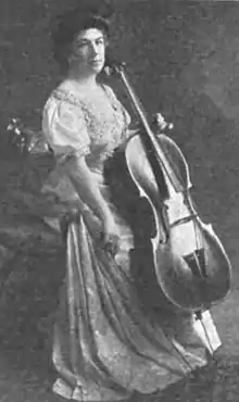 A middle-aged white woman wearing a gown, seated, holding a cello.