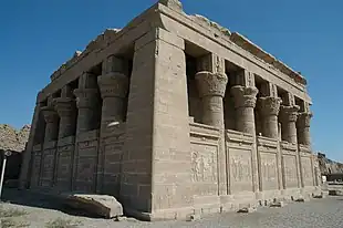 Small stone building surrounded by half-walls and columns with elaborate capitals