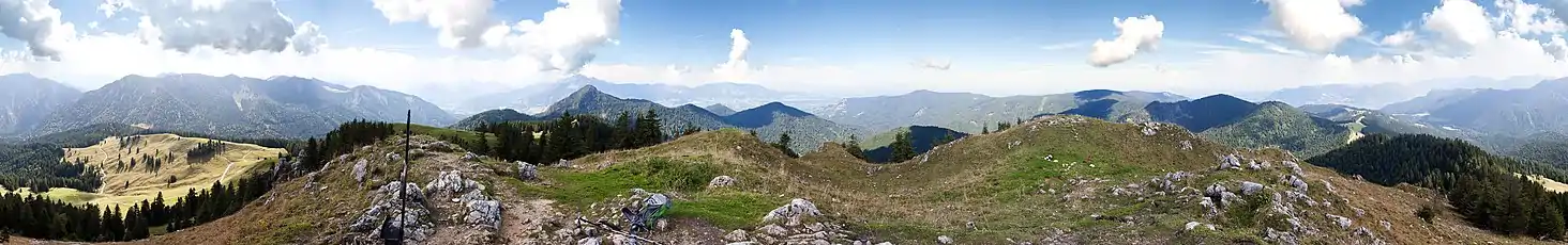 a 360 degree panorama from the top of a mountain. Scattered clouds are in the sky. The peak itself is grassy with some rocks jutting out, and the surrounding mountains are a mix of forests and meadows.