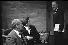 Three men in business suits, two sitting one standing, the standing one on the right is tall, slim, graying