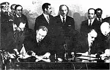 Two men signing an agreement, with other men standing behind them