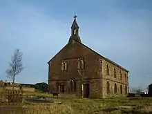 A two-storey stone church with round-headed windows and a bellcote on the near gable