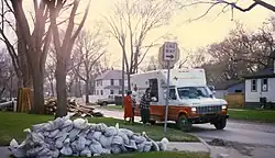 Several people are collecting items from an American Red Cross vehicle in a neighborhood. Around the neighborhood is piles of damaged possessions and sandbags.