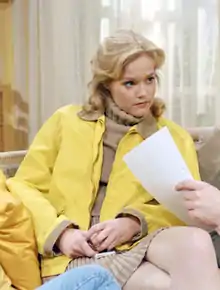 Guusje Nederhorst wearing a brown turtleneck sweater, brown skirt, and yellow jacket, sitting with legs crossed, looking at someone left of camera