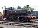 A black pannier tank locomotive, with the letters GWR in yellow on the side, pulls a mixed traffic train. The train includes a blue tank wagon, with "Express Dairy Milk for London" written on the side, and then a passenger carriage painted cream and red.