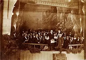 Orchestra in evening clothes assembled on a theatre stage, with a middle-aged white man with white hair and large white moustache standing in front of them, facing the camera