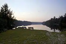  Grass and gravel lawn in the foreground, with a pair of benches on a shoreline in the center. In the background is a smooth lake surrounded by trees under a dusk sky.