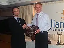 A man wearing a suit and a man wearing a shirt, tie and trousers, jointly holding a trophy.