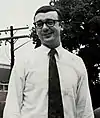 White male with dark hair and large dark glasses in a white shirt and dark tie